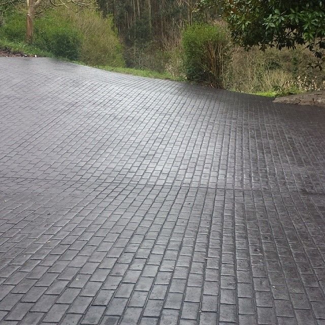 Entrada de garaje de una casa unifamiliar pavimentada con hormigón impreso gris textura adoquín y un coche aparcado.